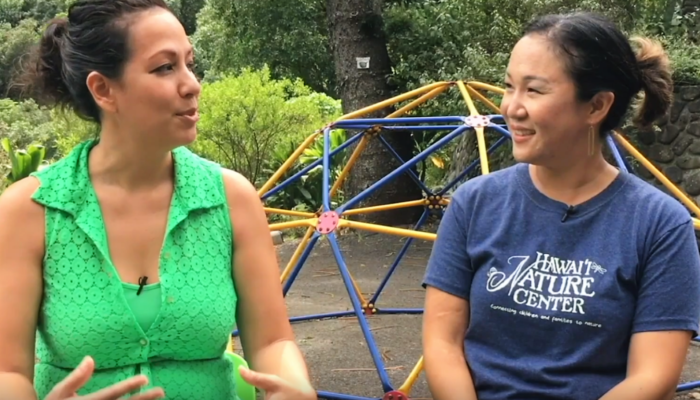 Two women sitting together, smiling, in front of a colorful playground structure on a sunny day.