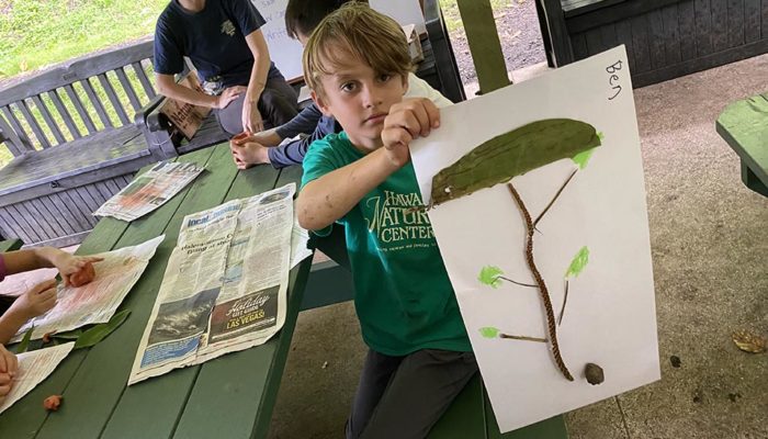 A young boy proudly displays a paper drawing of a tree using fallen leaves and branches.