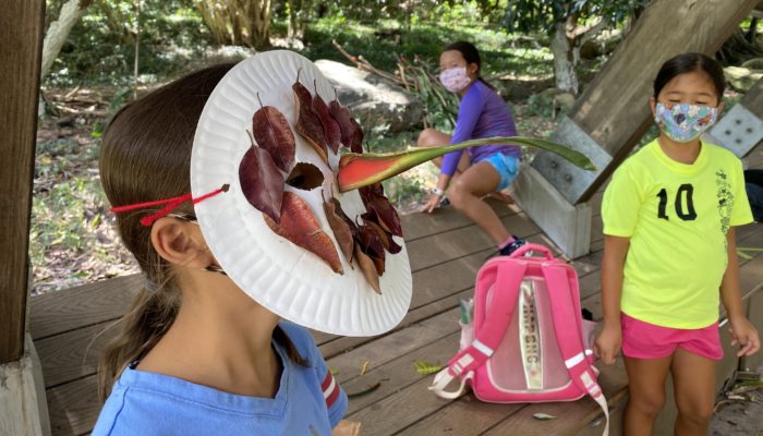 A young girl wearing a bird mask crafted from fallen leaves, yarn, and a paper plate, showcasing her creativity.